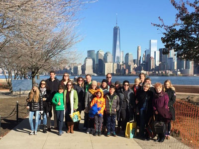 Group of visitors posing near the waterfront with the Lower Manhattan skyline and One World Trade Center behind them.